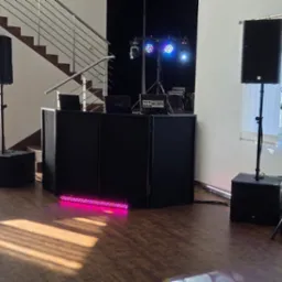 Profesjonalny zestaw nagłośnieniowy i oświetleniowy na parkiecie, gotowy do imprezy. Widoczne kolumny głośnikowe na statywach, konsoleta DJ-ska i efekty świetlne.