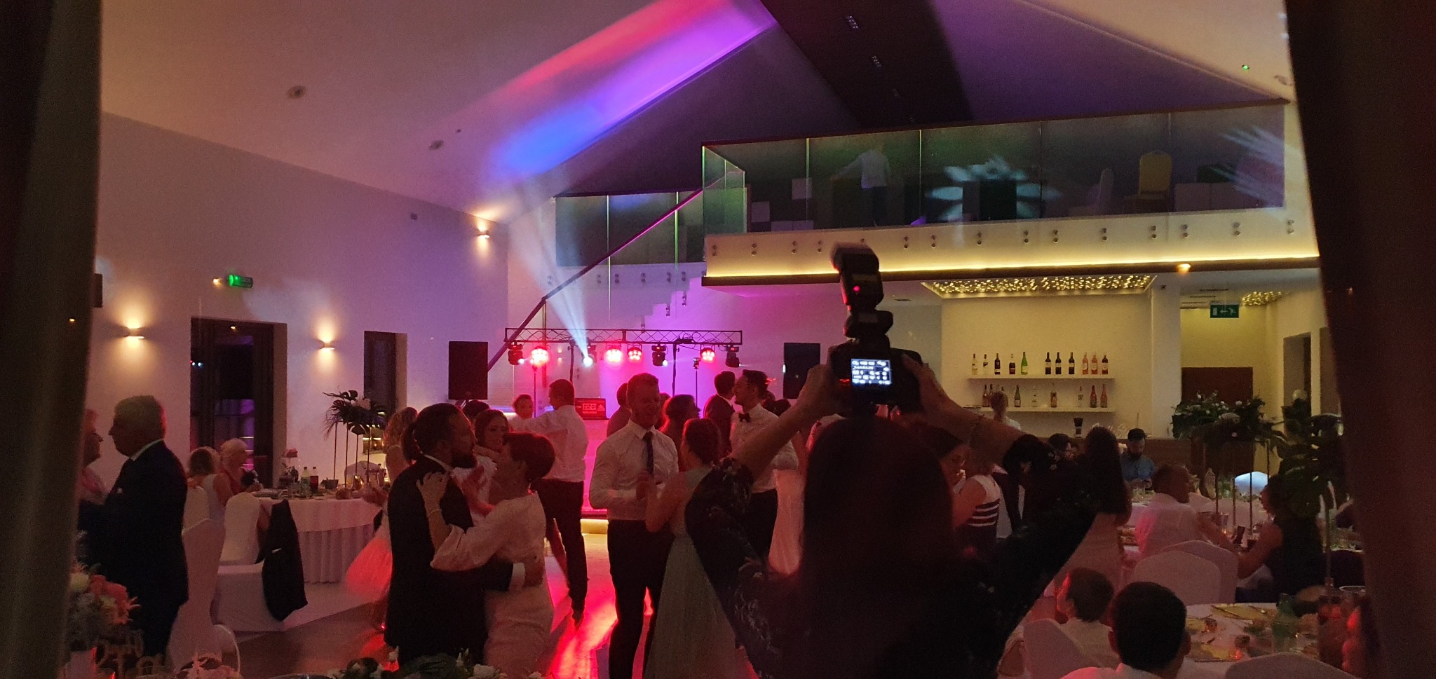 Sala weselna z parkietem, gdzie tańczą pary. W tle scena z oświetleniem i nagłośnieniem. W pierwszym planie fotograf robiący zdjęcie.