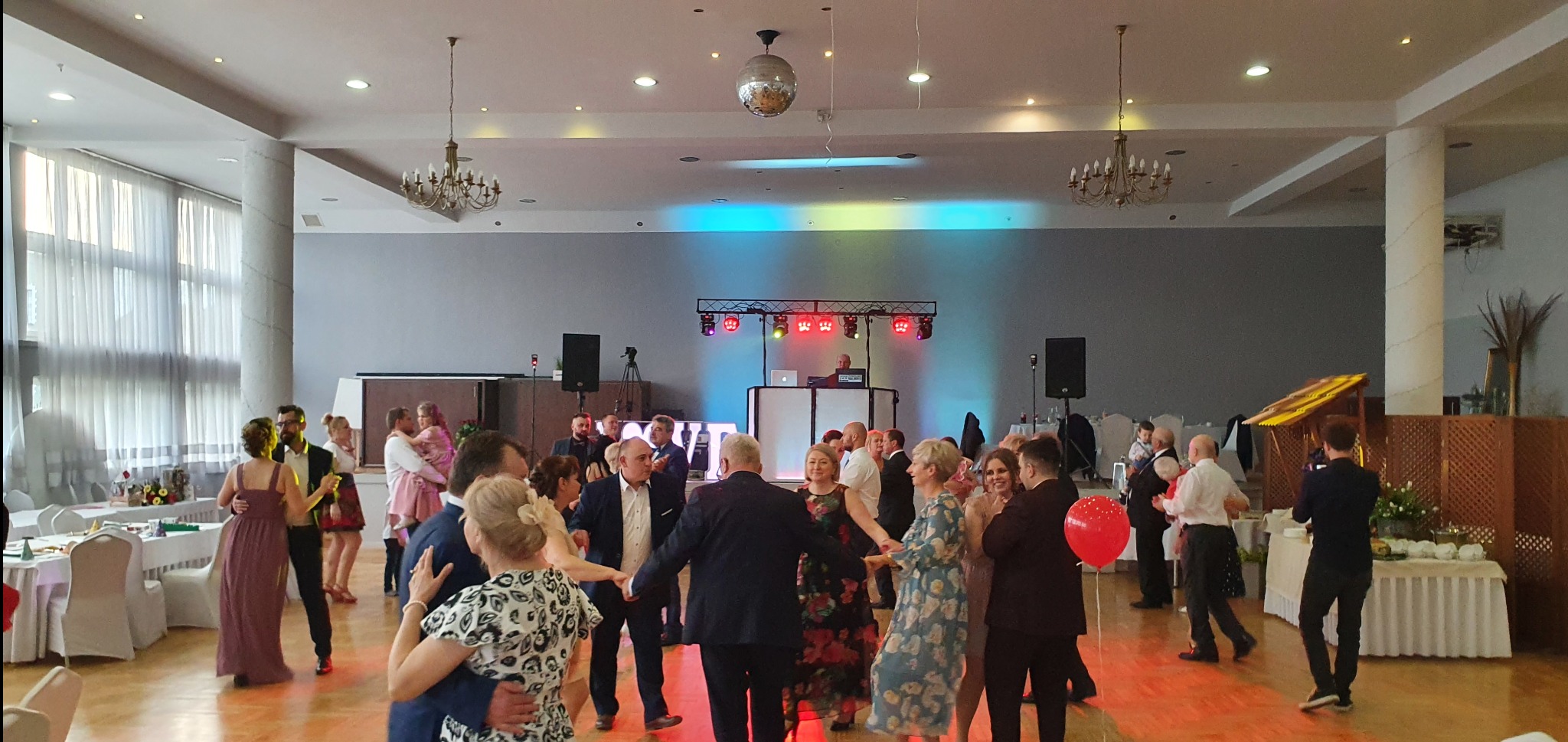 Sala balowa z tańczącymi gośćmi, dekoracjami, oświetleniem scenicznym i didżejem w tle, tworząca radosną atmosferę imprezy.