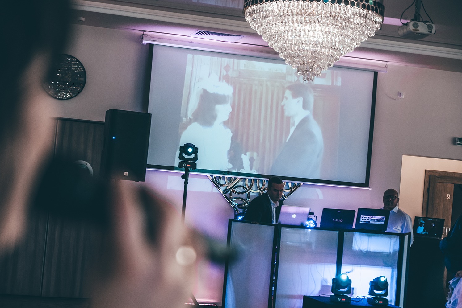 DJ na weselu wyświetla czarno-białe zdjęcie pary młodej na projektorze. W tle kryształowy żyrandol i głośniki. Scena oświetlona fioletowym światłem.