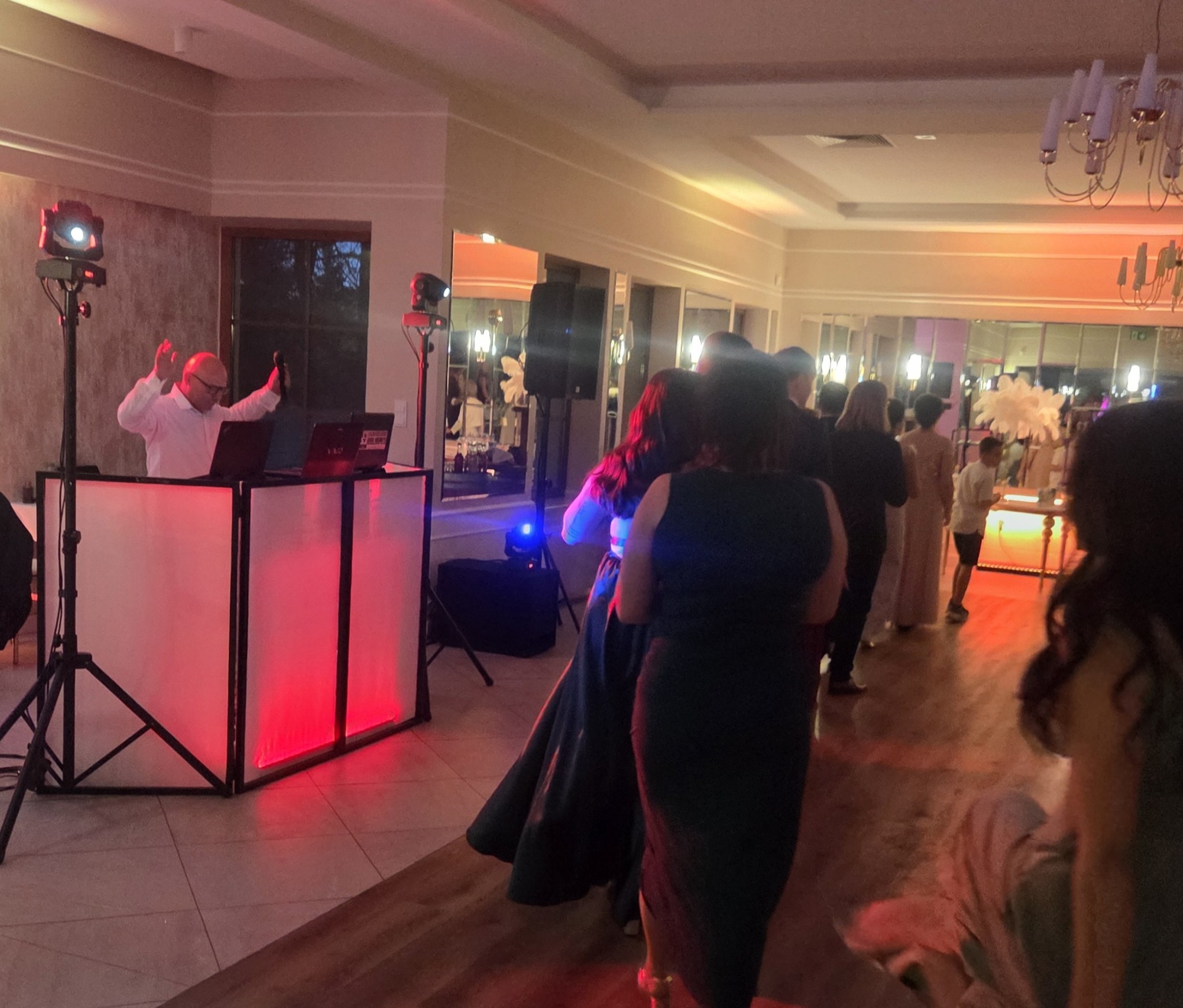 DJ z podświetlanym stanowiskiem gra muzykę na parkiecie podczas imprezy, w tle bawią się goście. Widoczny sprzęt nagłośnieniowy i oświetleniowy.