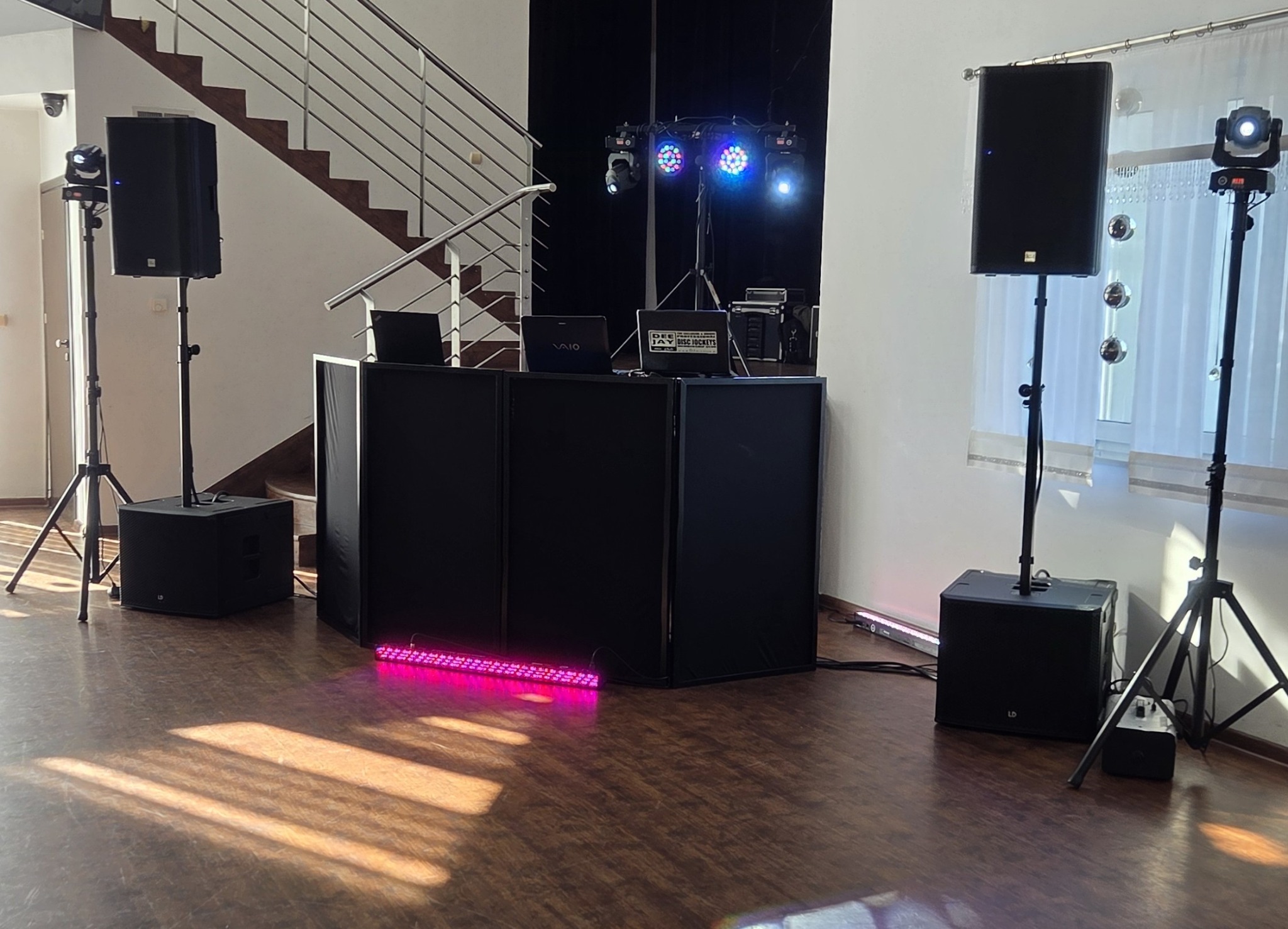 Profesjonalny zestaw nagłośnieniowy i oświetleniowy na parkiecie, gotowy do imprezy. Widoczne kolumny głośnikowe na statywach, konsoleta DJ-ska i efekty świetlne.