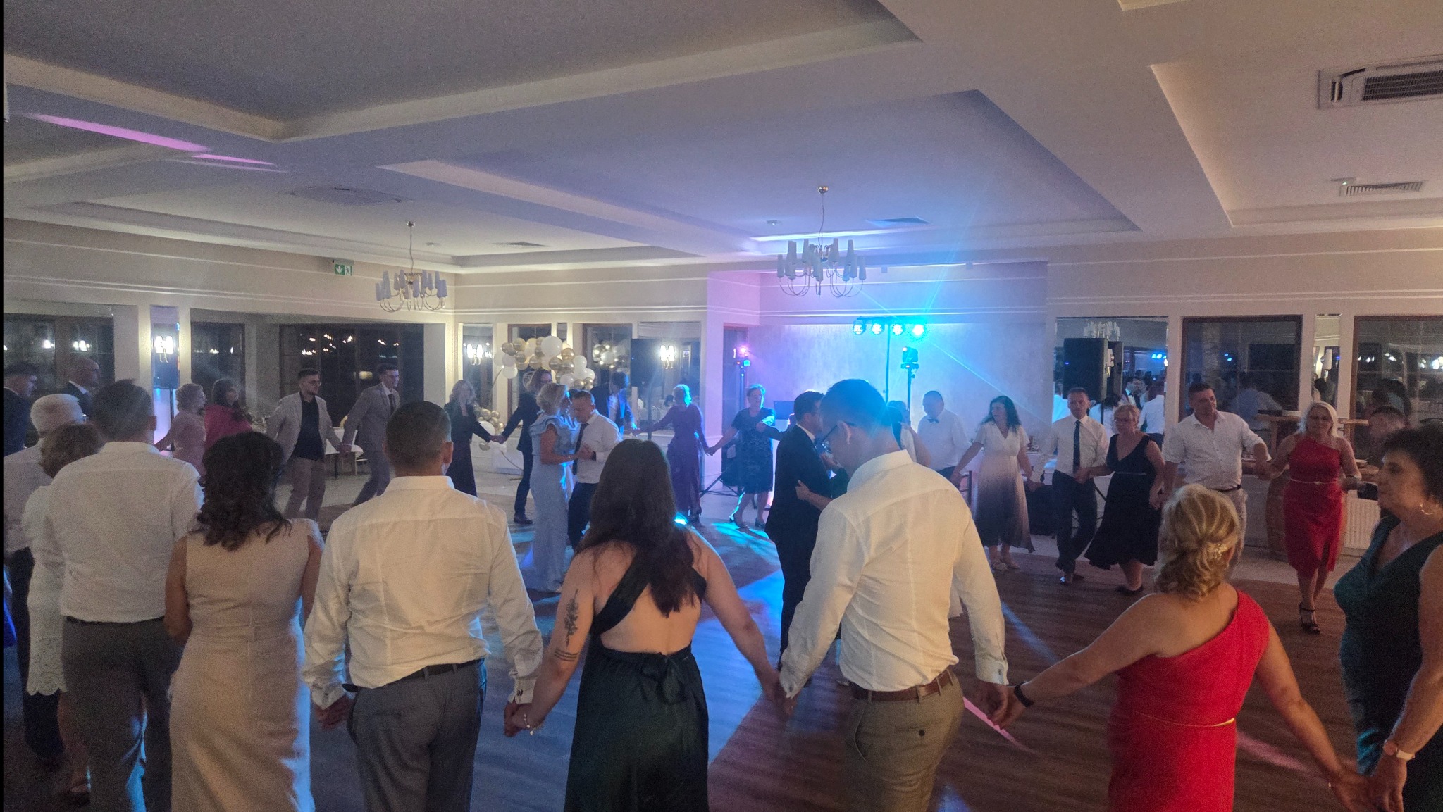 Goście weselni tańczą w kręgu, oświetleni niebieskimi światłami i z widocznymi dekoracjami sali. Uczestnicy w różnorodnych strojach, widok z tyłu.