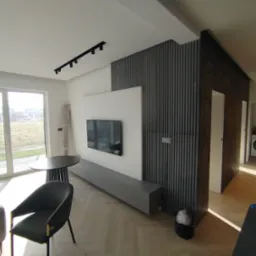 Nowoczesny salon z szarą sofą, stołem i krzesłem. Na ścianie telewizor i dekoracyjne panele pionowe. Jasne, przestronne wnętrze z parkietem.