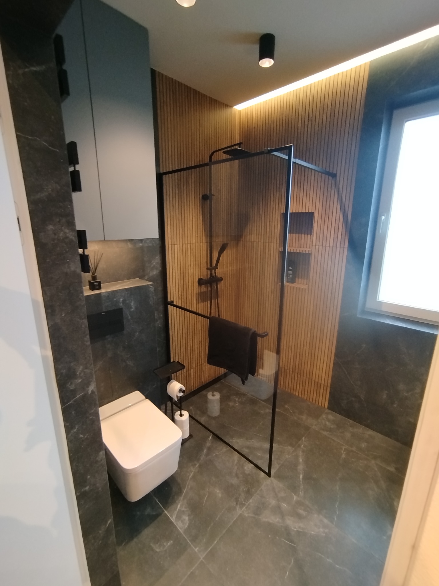 Nowoczesna łazienka z kabiną prysznicową typu walk-in, wykończona płytkami imitującymi marmur i drewnianymi panelami ściennymi. Minimalistyczny design, eleganckie oświetlenie.