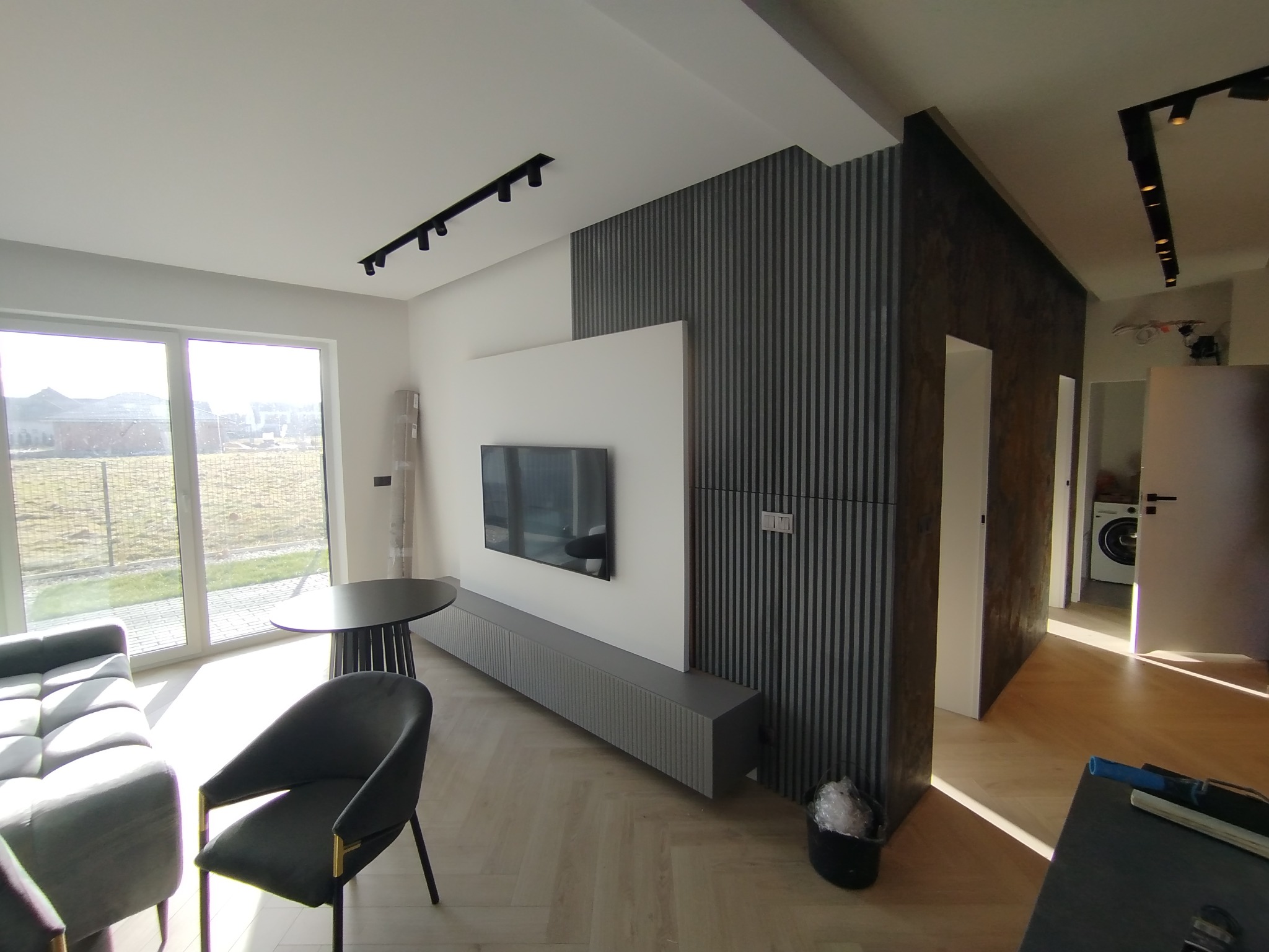 Nowoczesny salon z szarą sofą, stołem i krzesłem. Na ścianie telewizor i dekoracyjne panele pionowe. Jasne, przestronne wnętrze z parkietem.