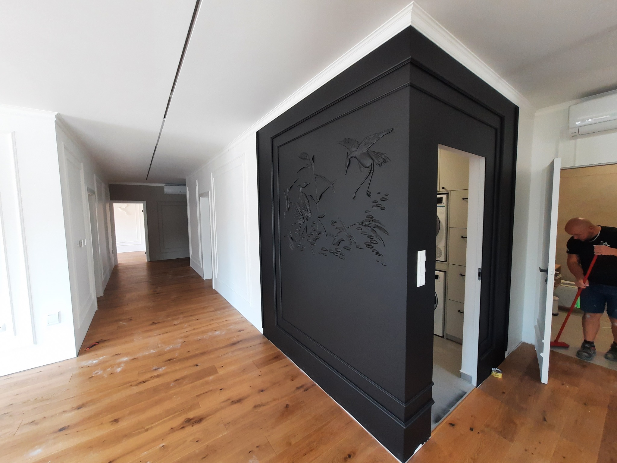 Elegancki korytarz po remoncie z drewnianą podłogą i czarną ścianą z płaskorzeźbą ptaków, sprzątany przez mężczyznę. Białe ściany z dekoracyjnymi listwami.