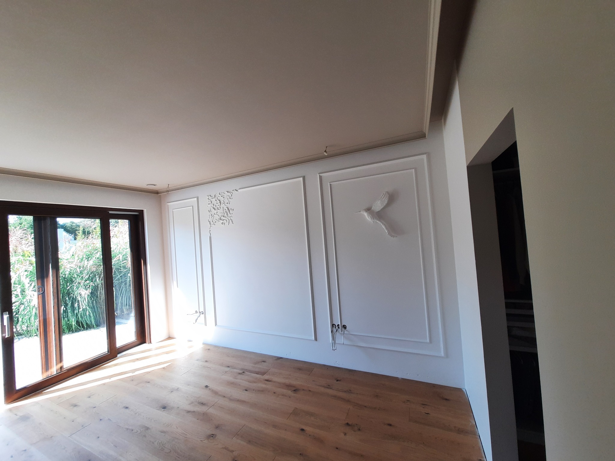 Eleganckie wnętrze po remoncie: białe ściany z dekoracyjnymi sztukateriami, motyw ptaka, drewniana podłoga i przesuwne drzwi balkonowe wpuszczające światło.