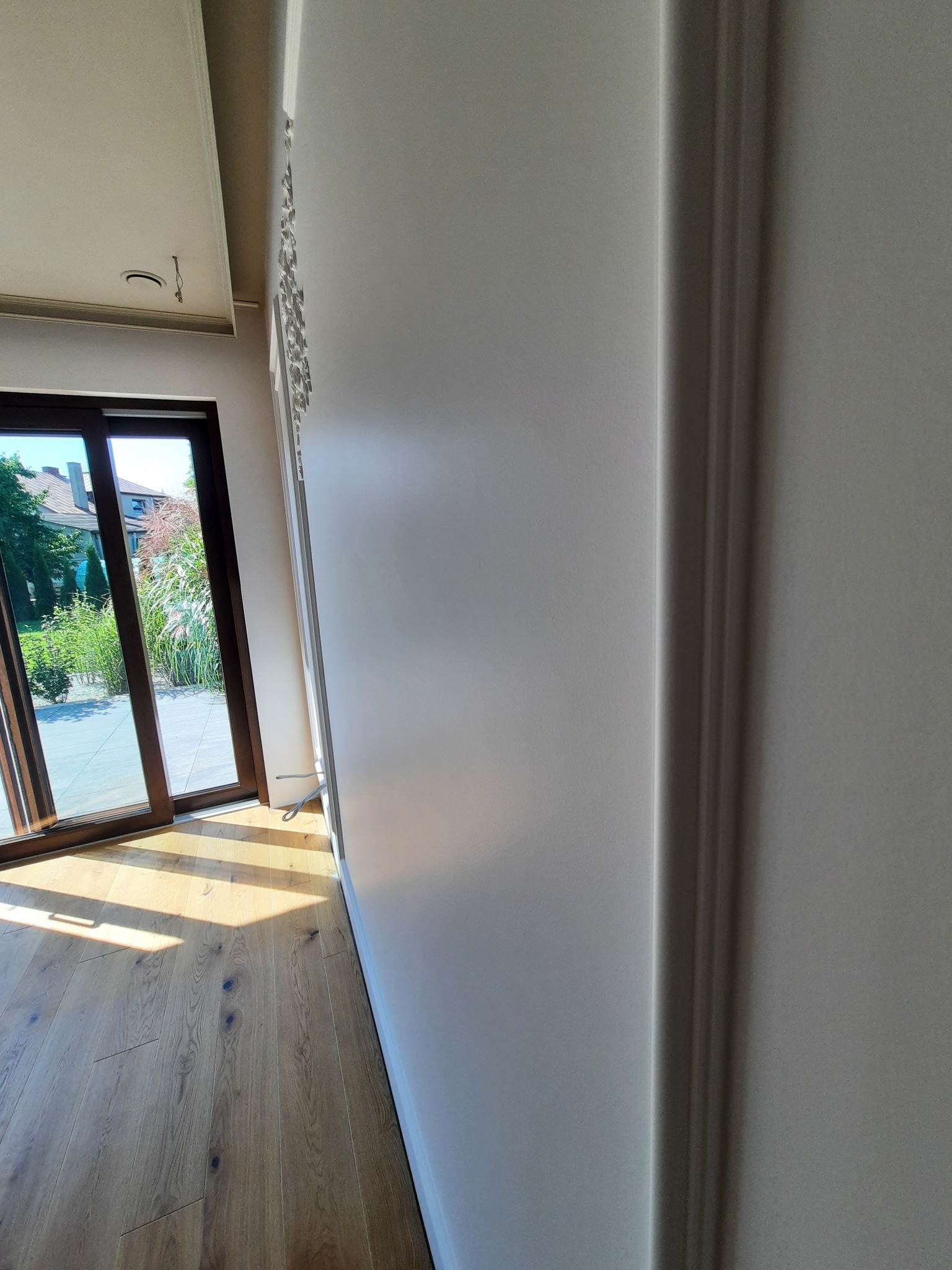 Jasny pokój po remoncie: świeżo pomalowane ściany z dekoracyjną sztukaterią, drewniana podłoga, widok na taras przez przesuwne drzwi.