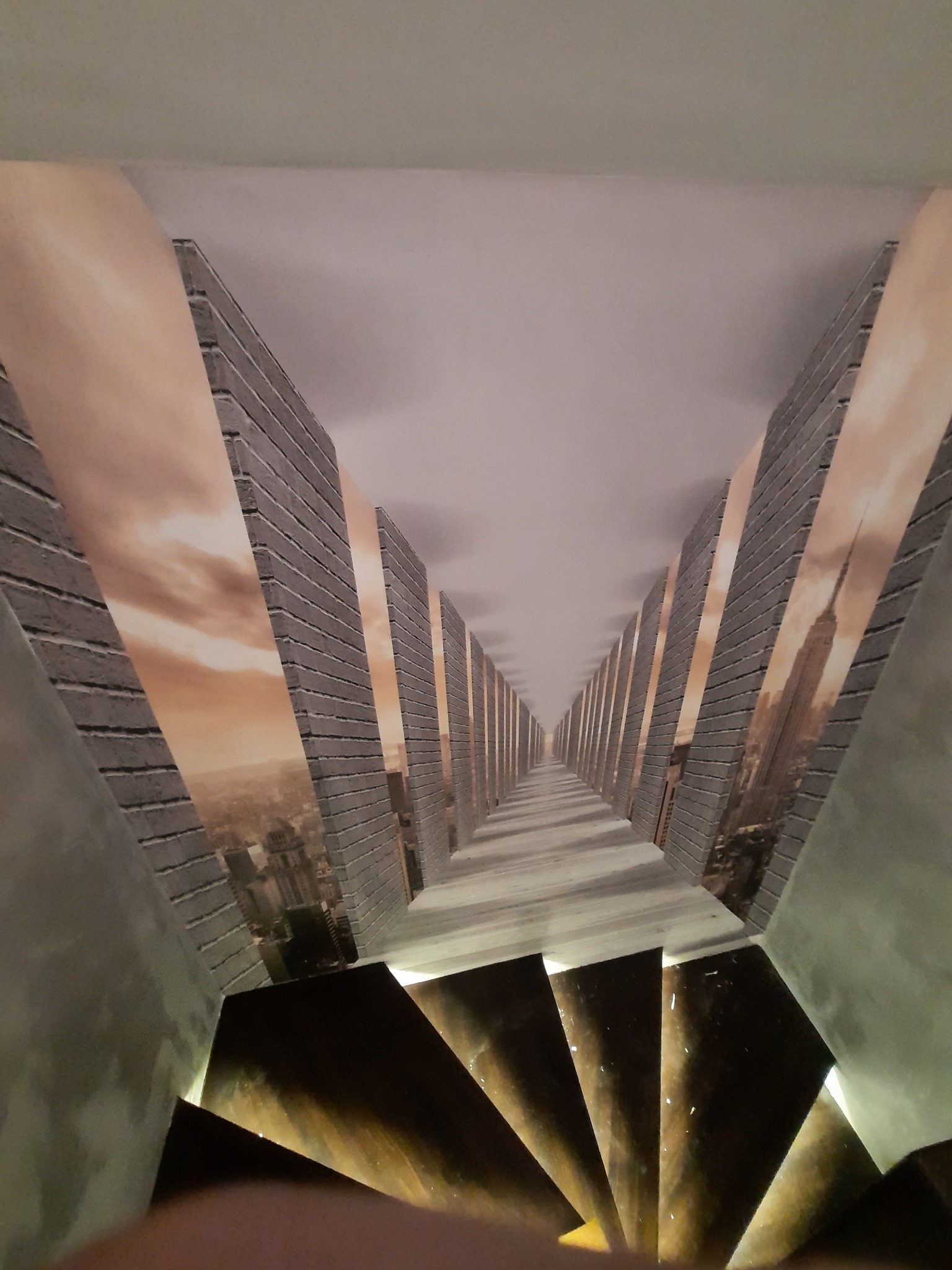 Fototapeta na ścianie w korytarzu, przedstawiająca perspektywę muru z widokiem na miasto, tworzy iluzję głębi. U dołu widoczne schody.