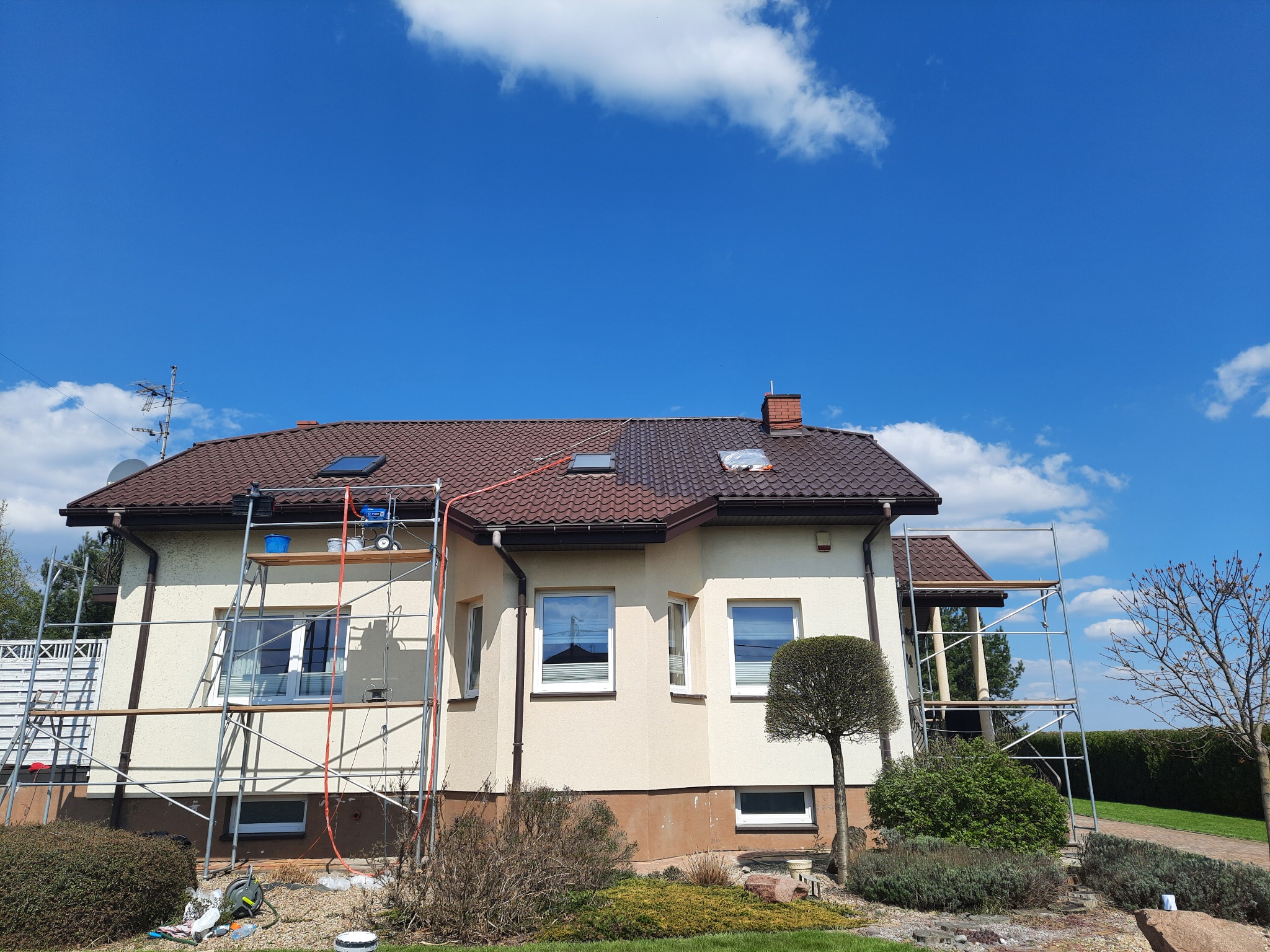 Dwukondygnacyjny dom z beżową elewacją w trakcie malowania. Rusztowania przy ścianach, błękitne niebo w tle. Widoczne narzędzia malarskie.