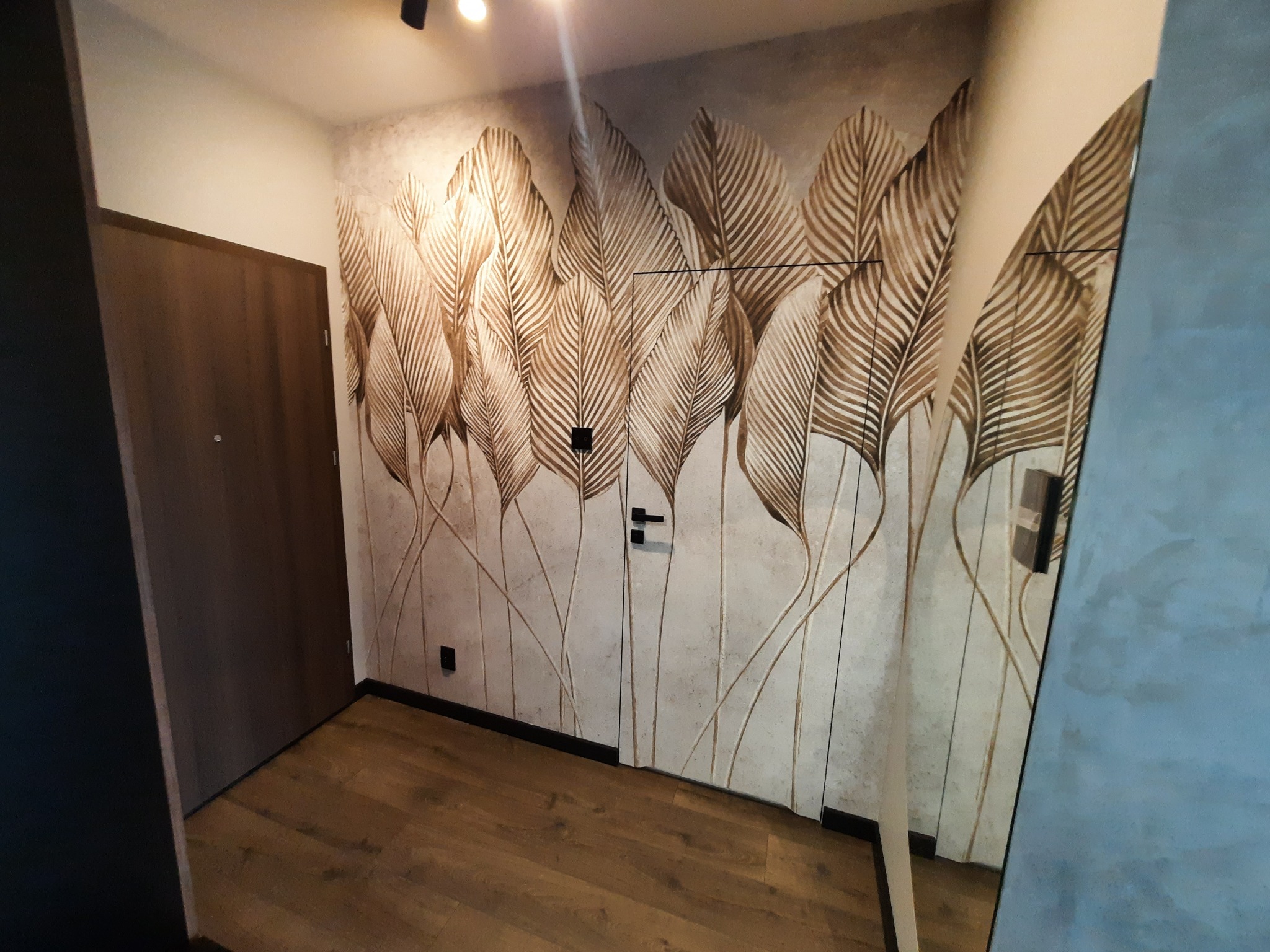 Nowoczesny przedpokój z drewnianą podłogą i tapetą w liście. Po prawej stronie lustro. Drzwi wejściowe w kolorze brązowym.