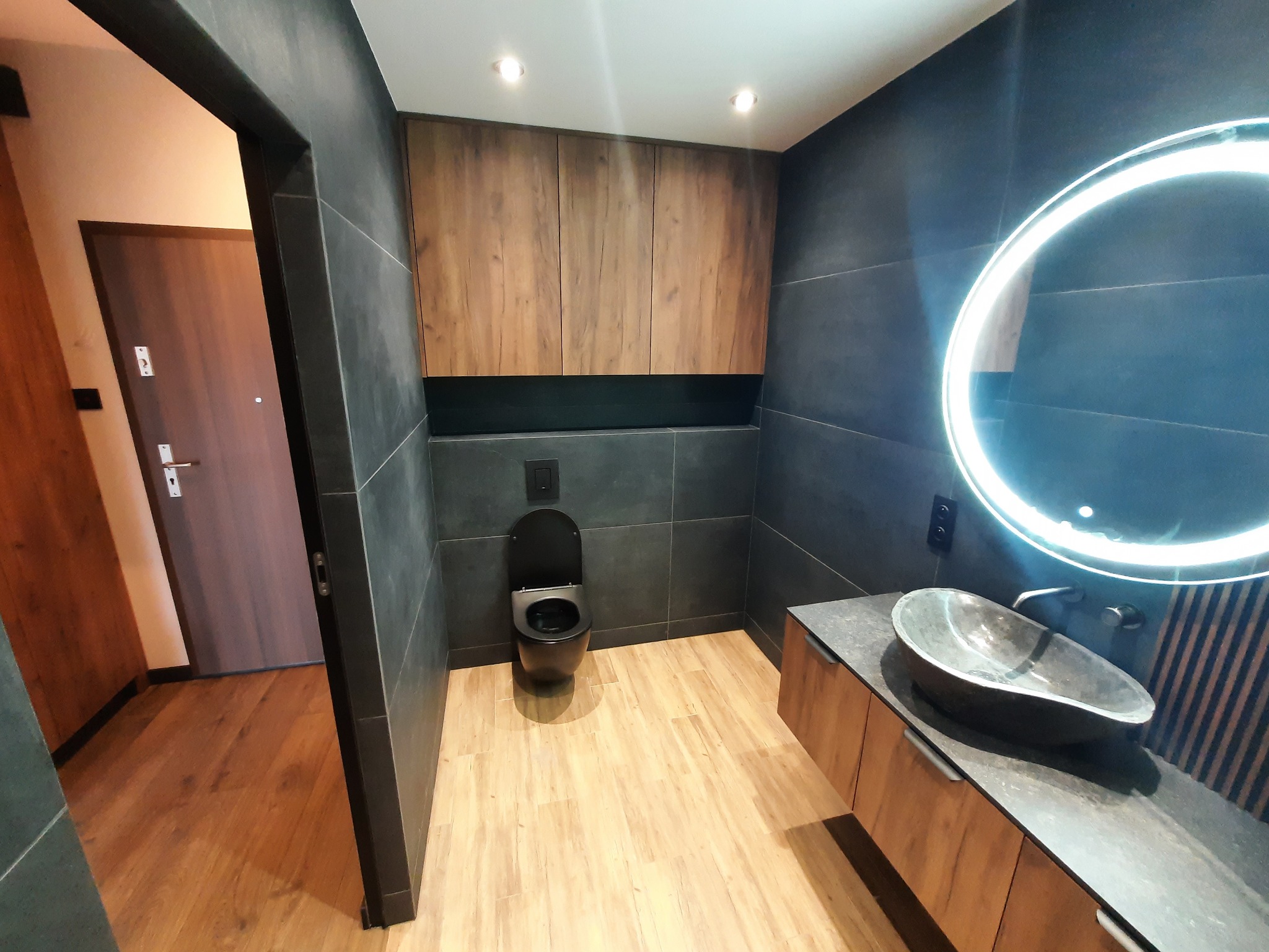 Nowoczesna łazienka z czarną armaturą, drewnianymi szafkami i okrągłym lustrem z podświetleniem LED. Minimalistyczny design, widok na podłogę i drzwi.