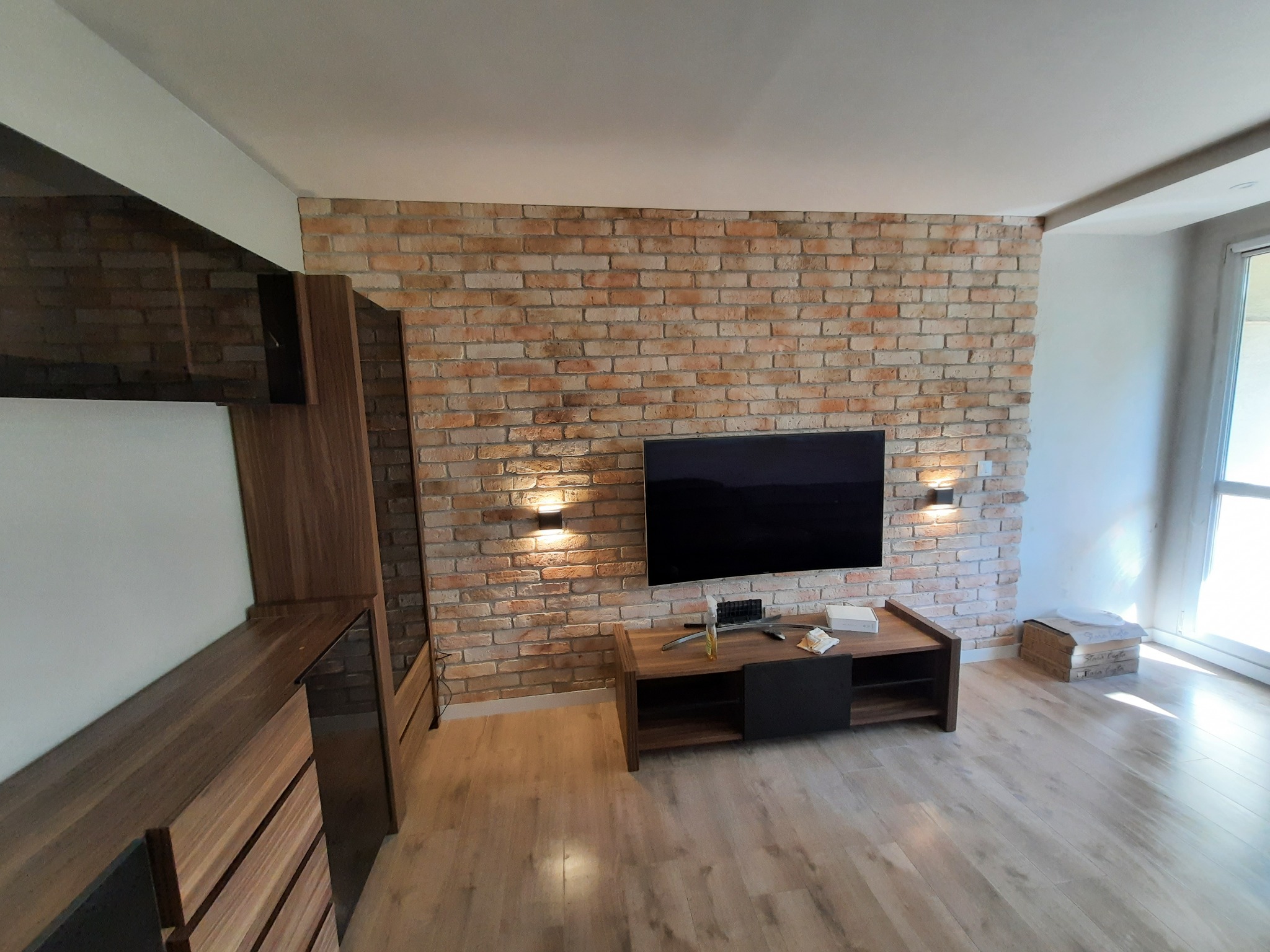 Salon z ceglaną ścianą i drewnianymi meblami. Telewizor wisi na ścianie, a pod nim stoi szafka RTV. Podłoga wyłożona panelami. Całość w ciepłych barwach.