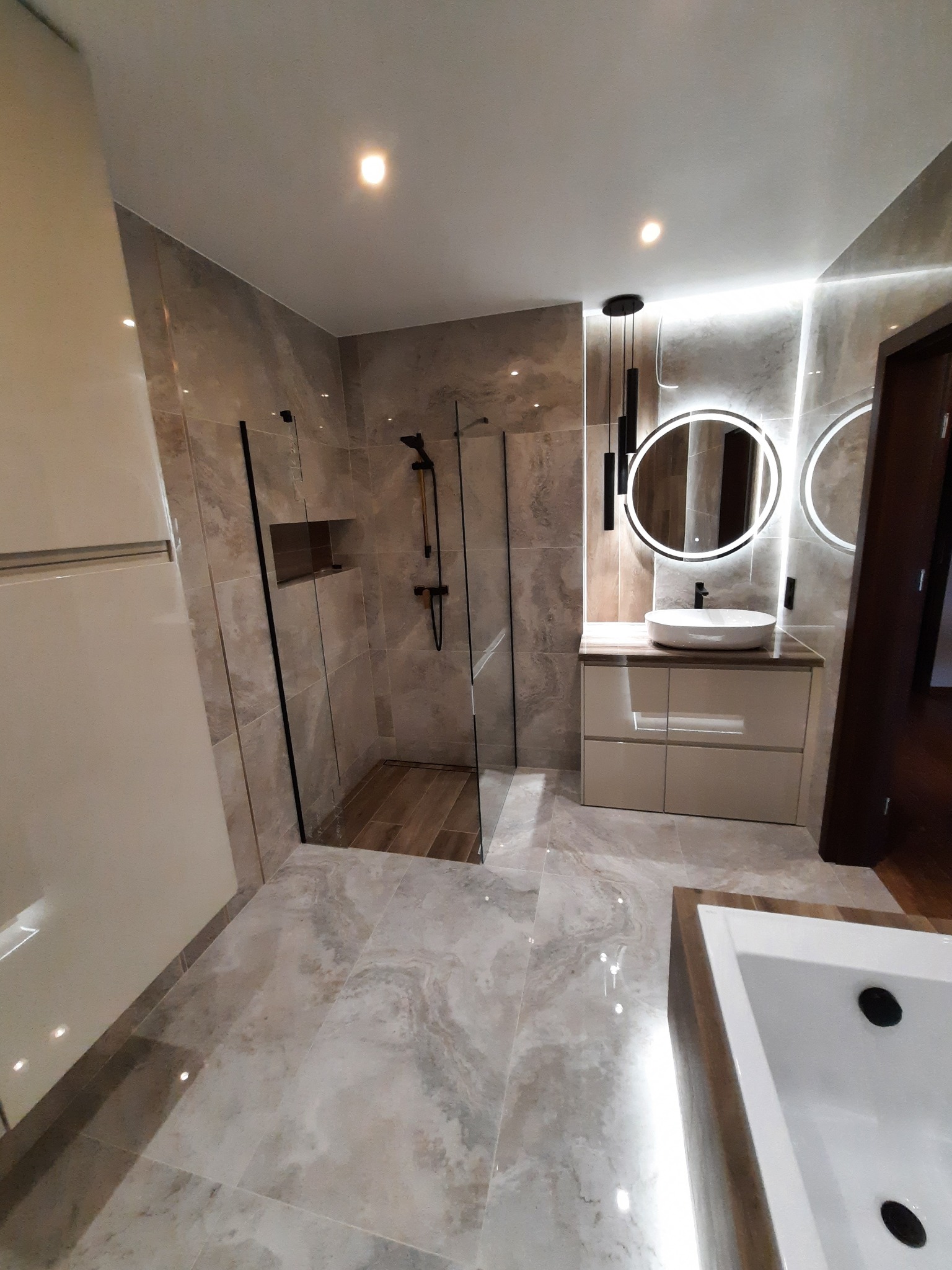 Nowoczesna łazienka z kabiną prysznicową typu walk-in, marmurowymi płytkami i podświetlanym lustrem. Minimalistyczny design z akcentami czerni.