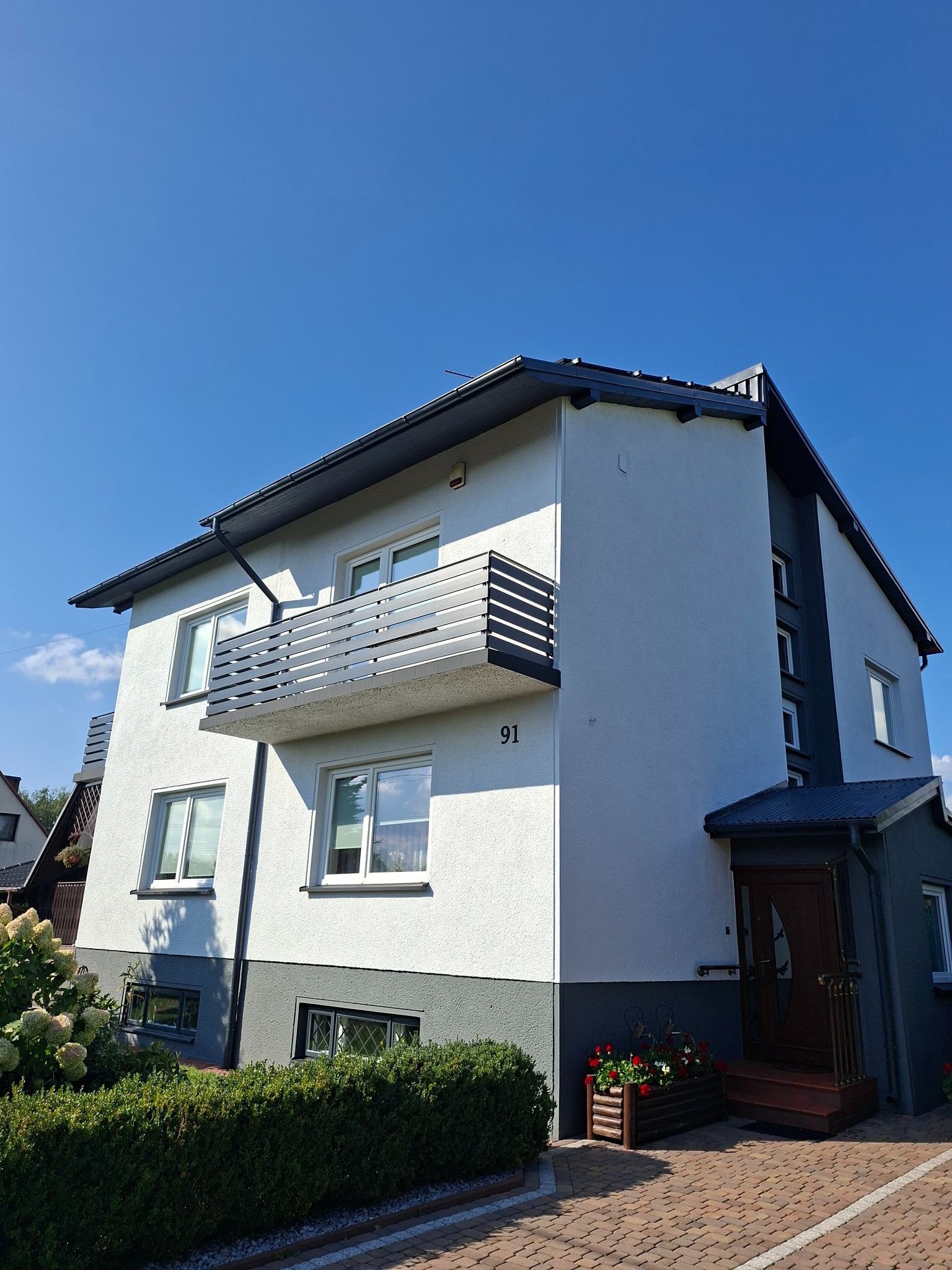 Odnowiona elewacja dwukondygnacyjnego domu z balkonem, numerem 91 i ciemnoszarą podmurówką. Widoczne zadbane krzewy i kwiaty w donicach.