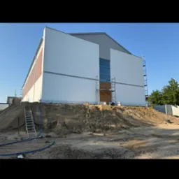 Ocieplony budynek z częściowo wykończoną elewacją, widoczne rusztowania i przygotowany teren. Fragment budynku z cegły. Jasne, słoneczne oświetlenie.