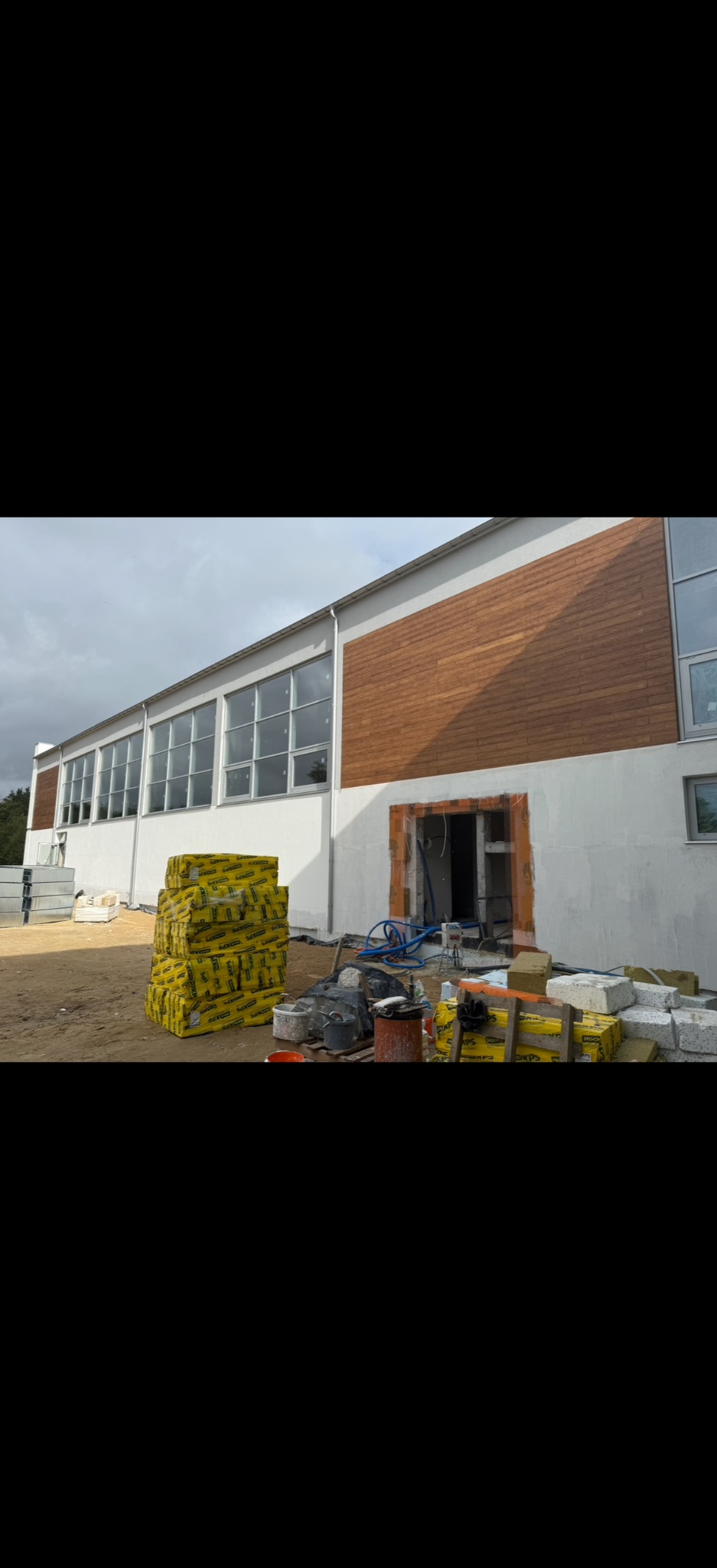 Budynek w trakcie prac elewacyjnych: częściowo ocieplony, z widocznym oknem i stertą materiałów izolacyjnych. Brązowe panele elewacyjne i białe ściany.