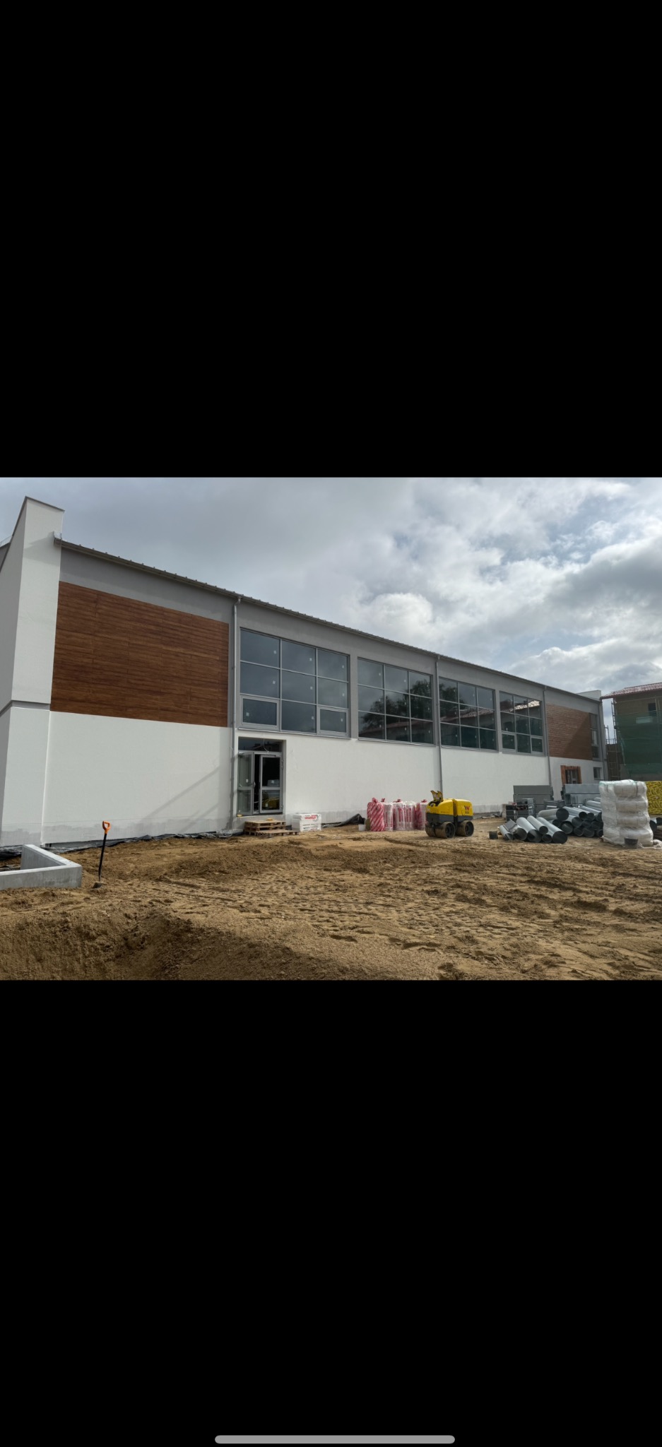 Budynek z białą elewacją i drewnianymi panelami, duże okna, teren budowy z piaskiem, ubijarka i materiały budowlane. Prace wykończeniowe.