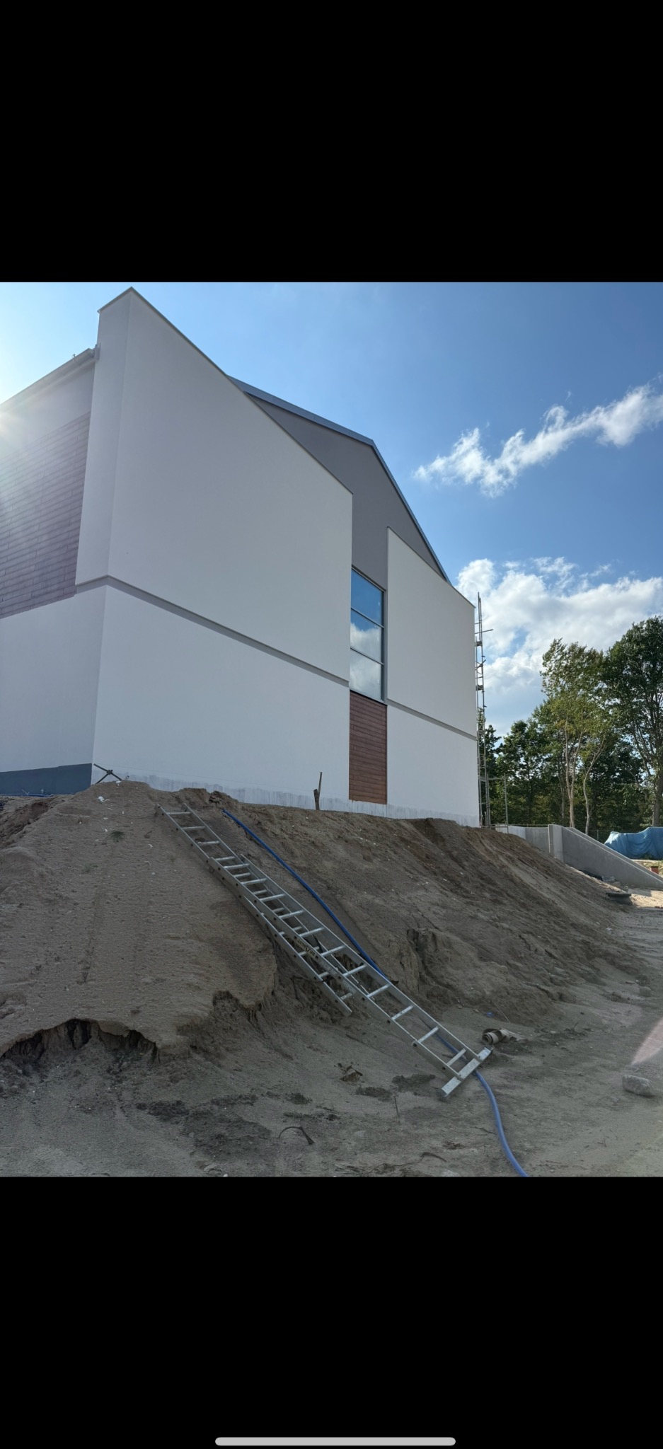 Nowoczesny dom z białą elewacją i szarym dachem w trakcie budowy. Na pierwszym planie ziemia i oparta o nią drabina. W tle drzewa i błękitne niebo.