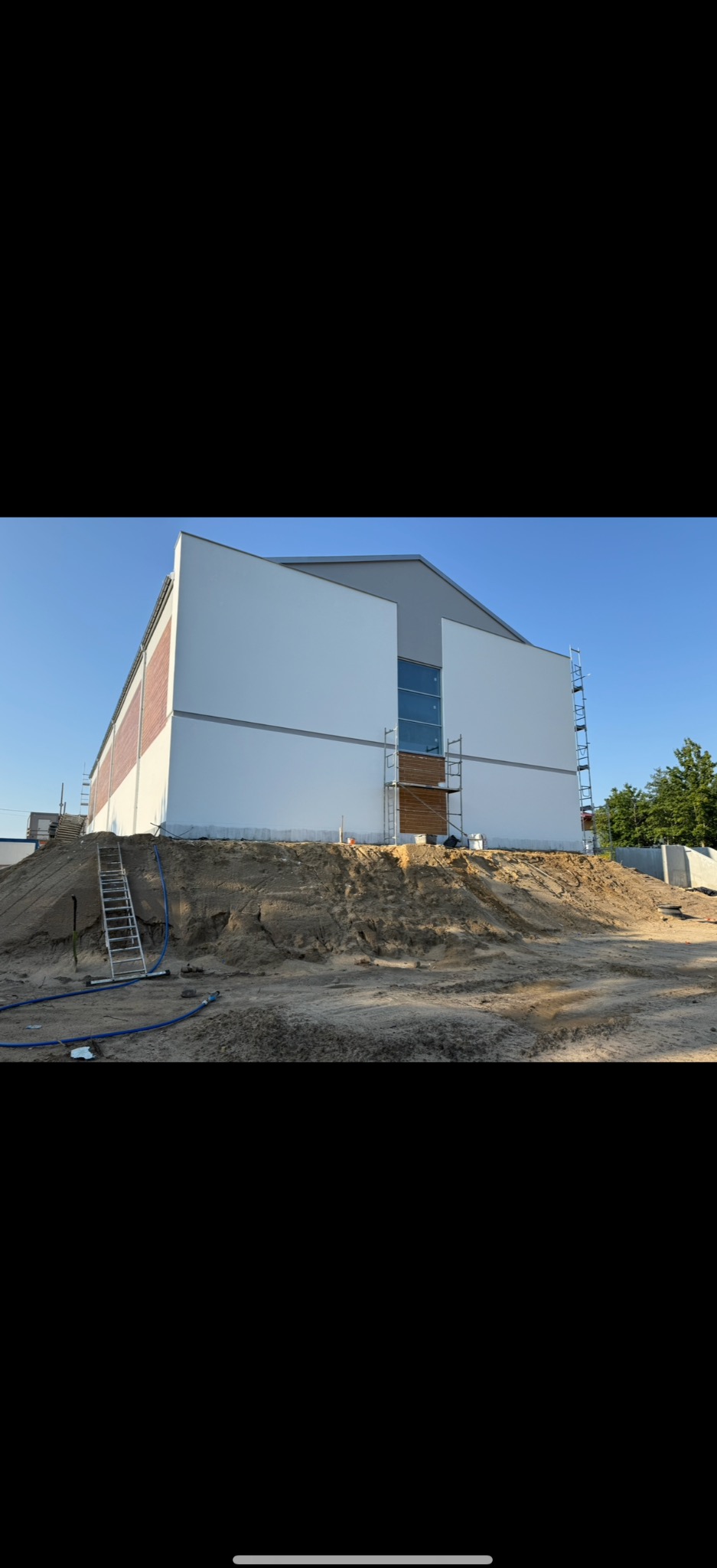 Ocieplony budynek z częściowo wykończoną elewacją, widoczne rusztowania i przygotowany teren. Fragment budynku z cegły. Jasne, słoneczne oświetlenie.