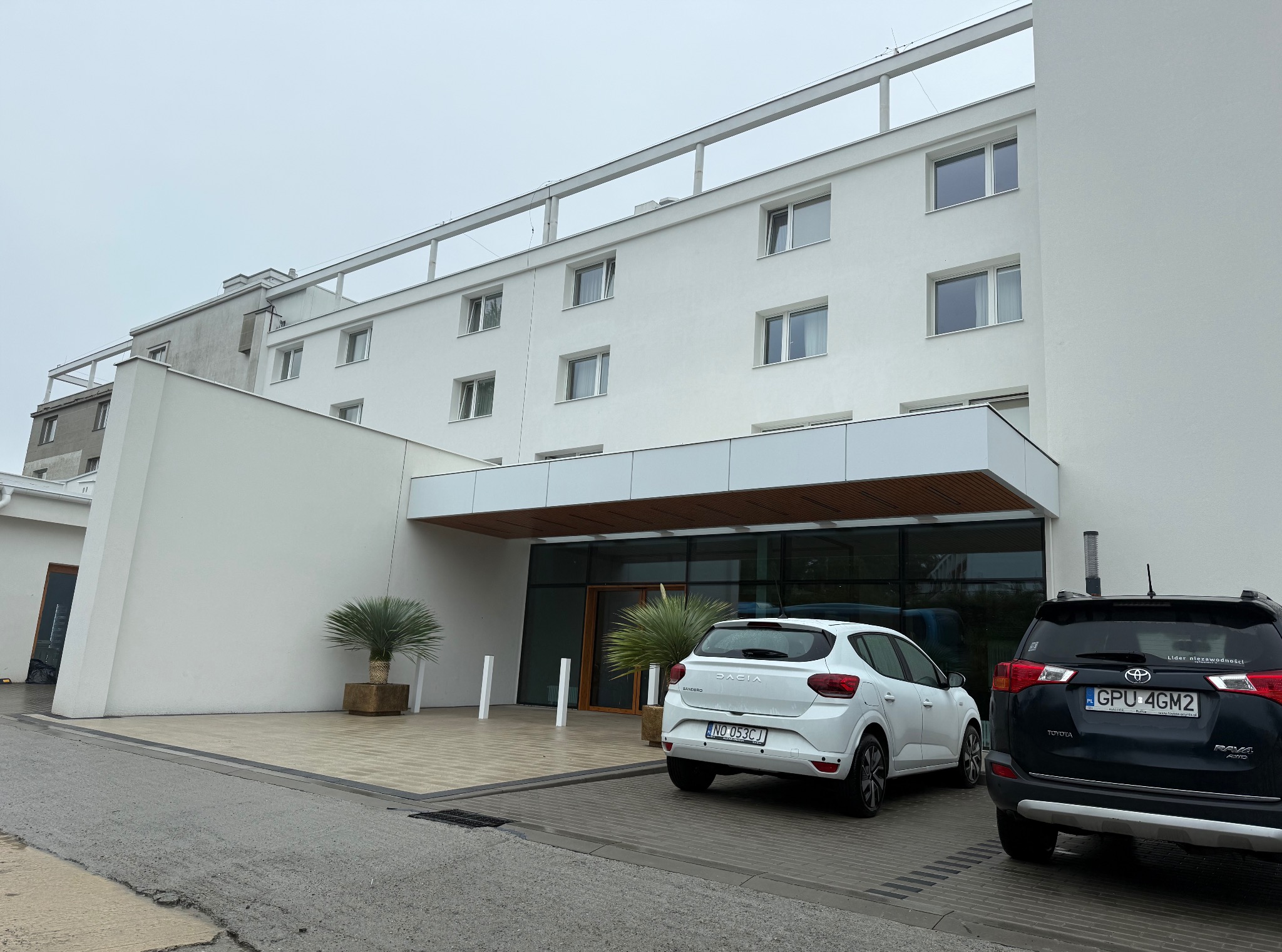 Nowoczesny, biały budynek z płaskim dachem i przeszklonym wejściem. Dwa samochody zaparkowane przed budynkiem. Minimalistyczna architektura.