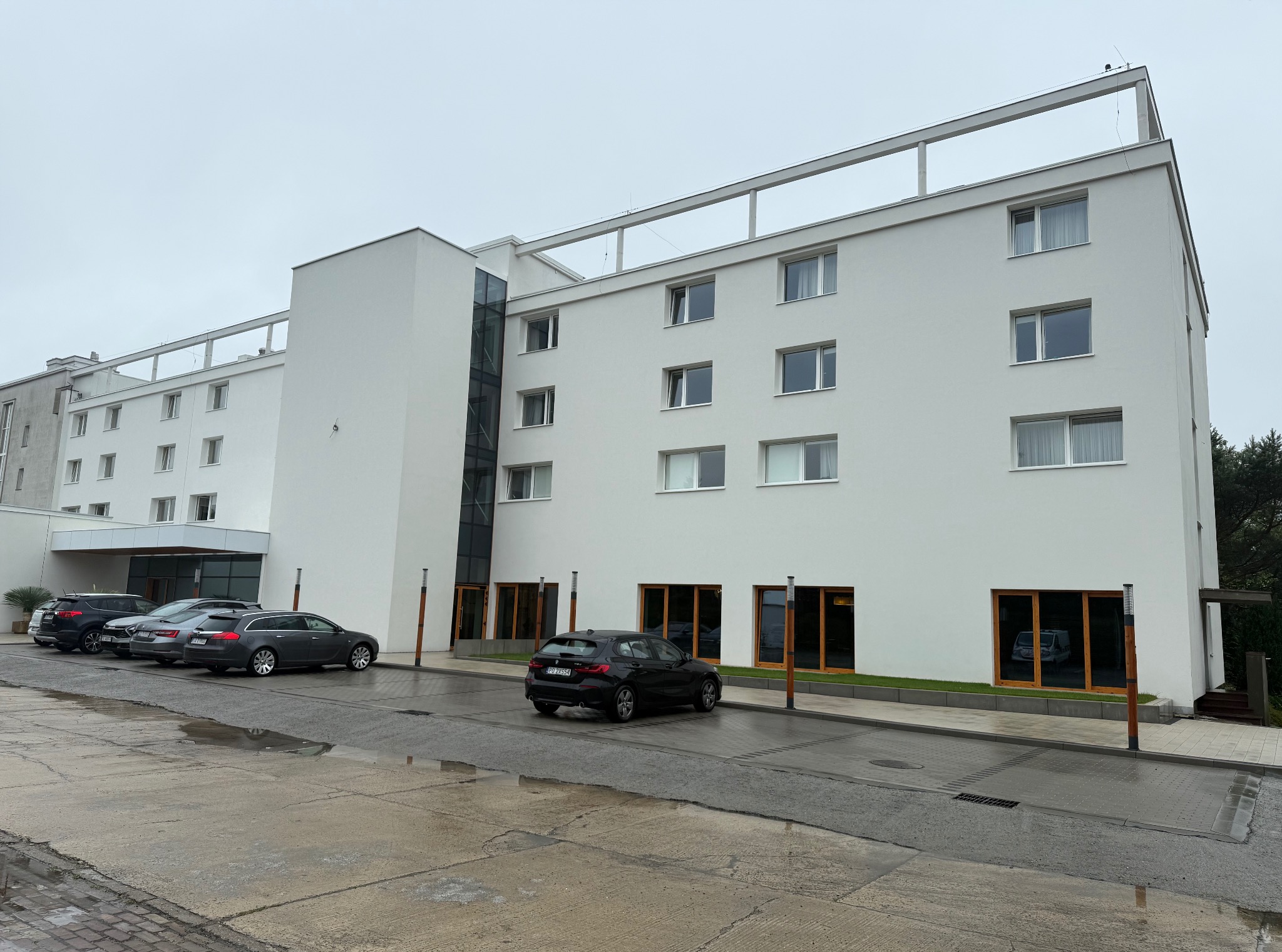 Biały budynek z płaskim dachem i jasnymi oknami. Na pierwszym planie zaparkowane samochody i mokry chodnik. Widoczne elementy elewacji i otoczenia.