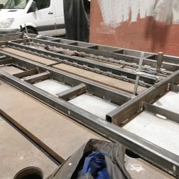 KB Krzysztof Bram - Konstrukcja metalowa z profili spawanych, widoczne spawy, w tle biały bus, elementy mocujące. Praca ślusarska w warsztacie, przygotowanie pod konstrukcję.