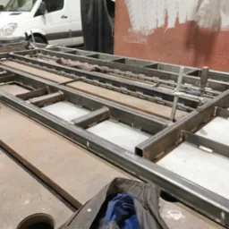 Konstrukcja metalowa z profili spawanych, widoczne spawy, w tle biały bus, elementy mocujące. Praca ślusarska w warsztacie, przygotowanie pod konstrukcję.