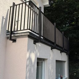 KB Krzysztof Bram - Czarna balustrada balkonowa z pionowymi szczeblami zamontowana na elewacji budynku z oknami i widocznymi elementami mocującymi. Tło stanowi zieleń drzew.