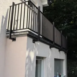 Czarna balustrada balkonowa z pionowymi szczeblami zamontowana na elewacji budynku z oknami i widocznymi elementami mocującymi. Tło stanowi zieleń drzew.