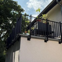 KB Krzysztof Bram - Czarna, metalowa balustrada balkonowa z widocznymi mocowaniami do ściany, na tle błękitnego nieba i zieleni drzew. Na balkonie donica z rośliną. Widok z dołu.