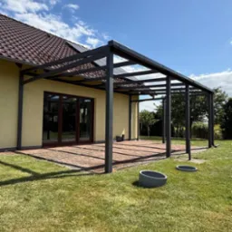 Nowoczesna pergola z ciemnego metalu z przeszklonym dachem, przylegająca do jasnego domu z brązowym dachem, na tle zielonego trawnika i błękitnego nieba.