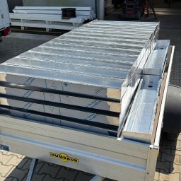 KB Krzysztof Bram - Transport przyczepą elementów konstrukcyjnych z blachy aluminiowej, widoczne zabezpieczenia folią ochronną. W tle brama hali i podnośnik koszowy.