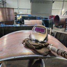 KB Krzysztof Bram - Hełm spawalniczy i szlifierka na metalowej konstrukcji w hali produkcyjnej. W tle widoczny spawacz przy pracy i elementy konstrukcji stalowych.