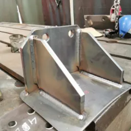 Metalowa konstrukcja spawana z otworami, widoczne spawy, leżąca na stole warsztatowym, w tle narzędzia i akcesoria spawalnicze. Detal konstrukcyjny.