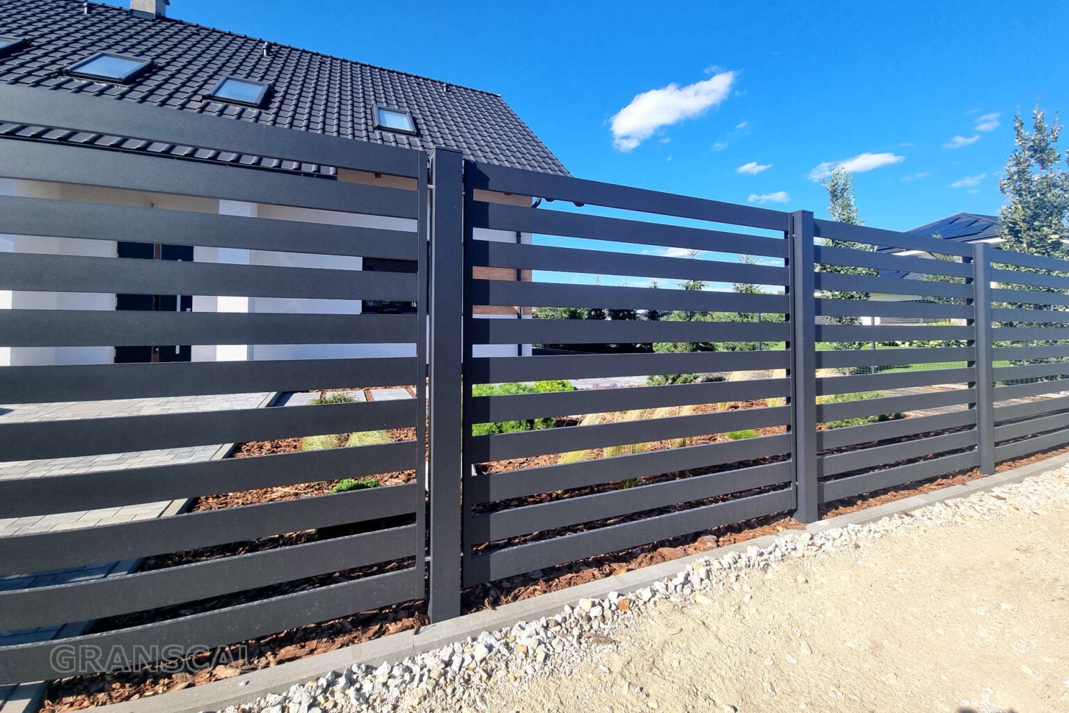 Nowoczesne, poziome ogrodzenie metalowe w kolorze antracytowym, z widocznym fragmentem domu i błękitnym niebem w tle. Minimalistyczny design, schludne wykonanie.