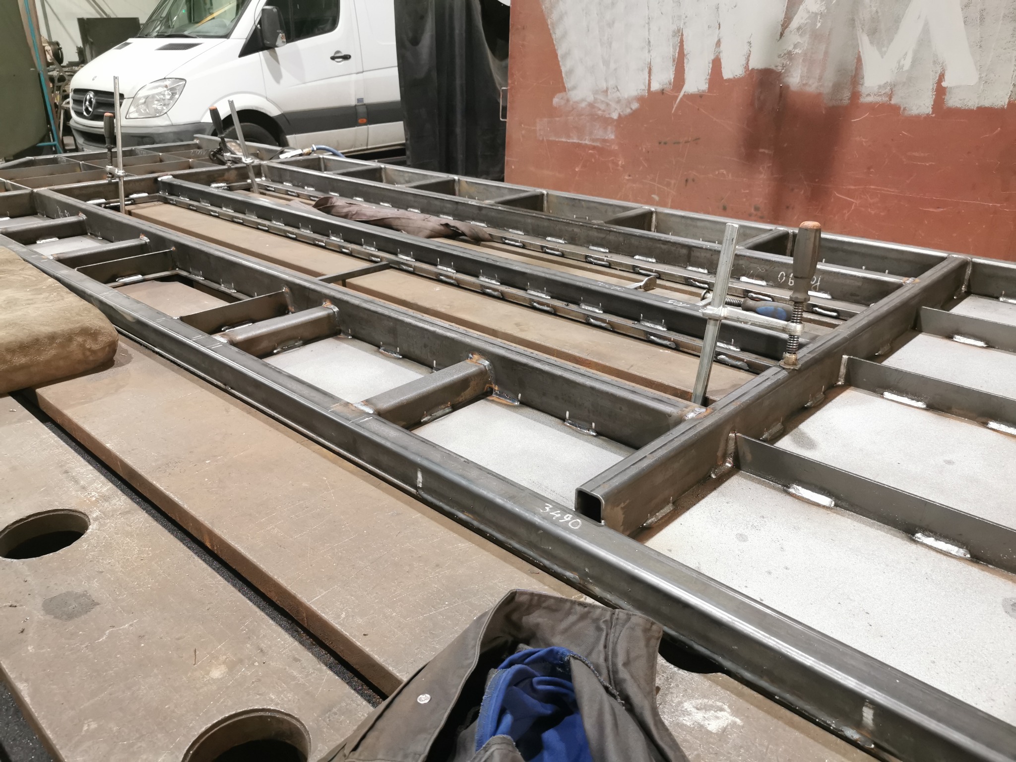 Konstrukcja metalowa z profili spawanych, widoczne spawy, w tle biały bus, elementy mocujące. Praca ślusarska w warsztacie, przygotowanie pod konstrukcję.