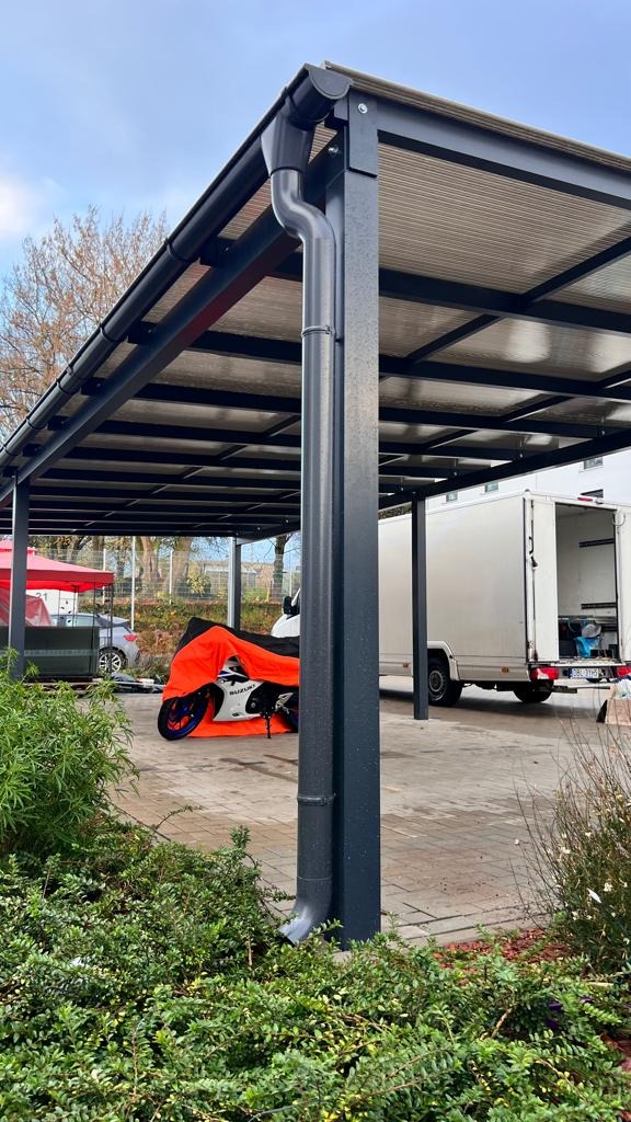 Nowoczesna, metalowa pergola z widocznym systemem odprowadzania wody deszczowej. Pod zadaszeniem zaparkowany motocykl przykryty pomarańczowym pokrowcem.