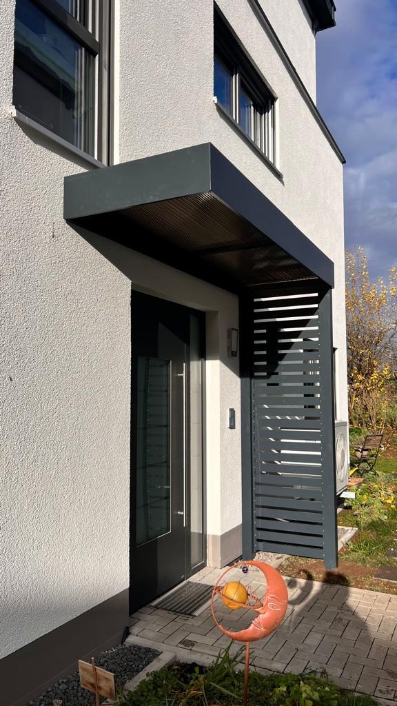 Nowoczesne zadaszenie nad wejściem do domu z metalową osłoną boczną z poziomych listew, widok z zewnątrz. Dekoracja w postaci metalowego księżyca.