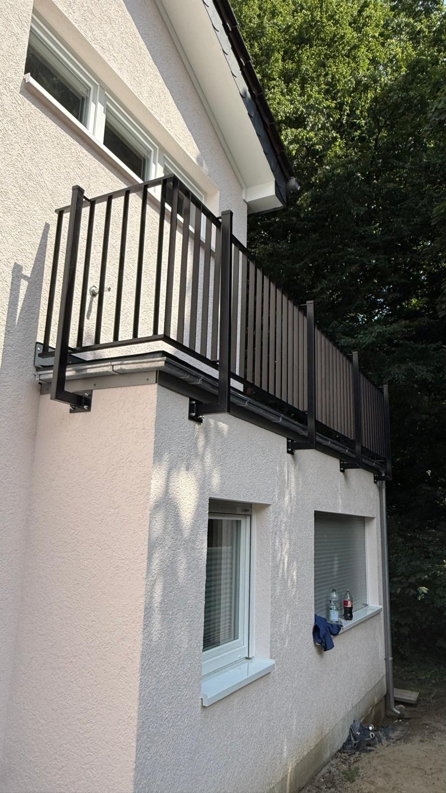 Czarna balustrada balkonowa z pionowymi szczeblami zamontowana na elewacji budynku z oknami i widocznymi elementami mocującymi. Tło stanowi zieleń drzew.