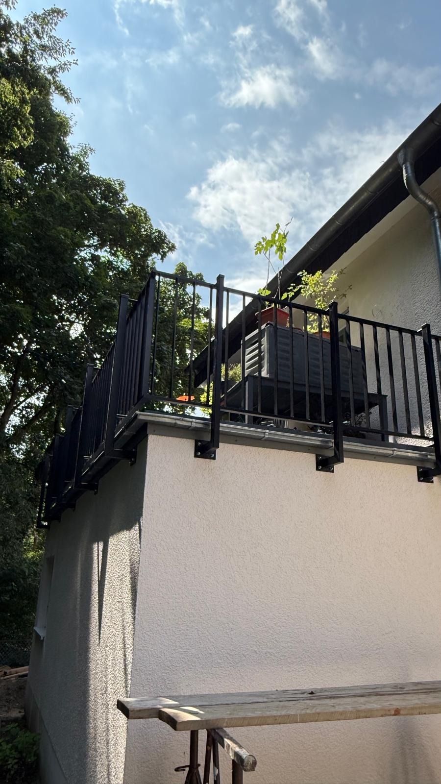 Czarna, metalowa balustrada balkonowa z widocznymi mocowaniami do ściany, na tle błękitnego nieba i zieleni drzew. Na balkonie donica z rośliną. Widok z dołu.