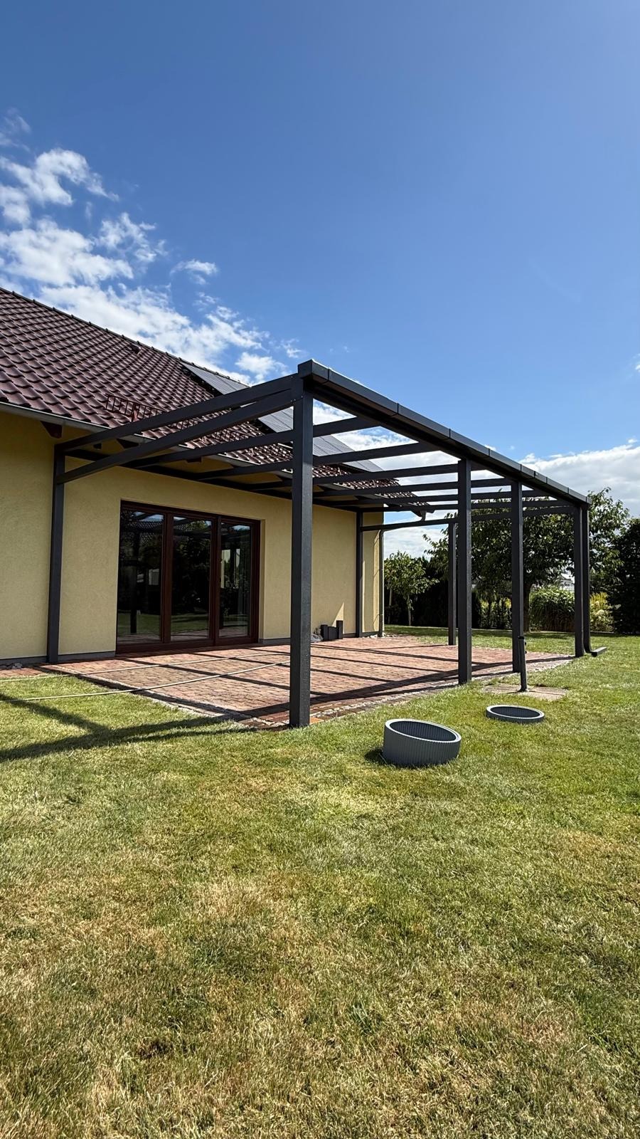Nowoczesna pergola z ciemnego metalu z przeszklonym dachem, przylegająca do jasnego domu z brązowym dachem, na tle zielonego trawnika i błękitnego nieba.