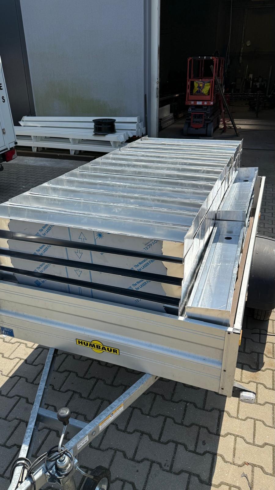 Transport przyczepą elementów konstrukcyjnych z blachy aluminiowej, widoczne zabezpieczenia folią ochronną. W tle brama hali i podnośnik koszowy.