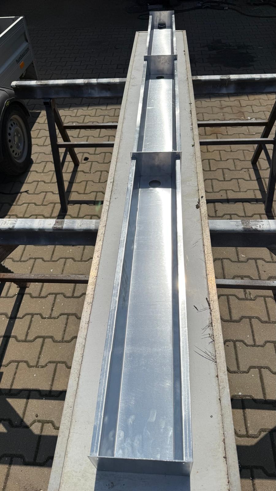 Długa, metalowa konstrukcja kanałowa z otworami, umieszczona na metalowym stelażu, widziana z góry na tle brukowanej nawierzchni. Element konstrukcyjny w surowym stanie.