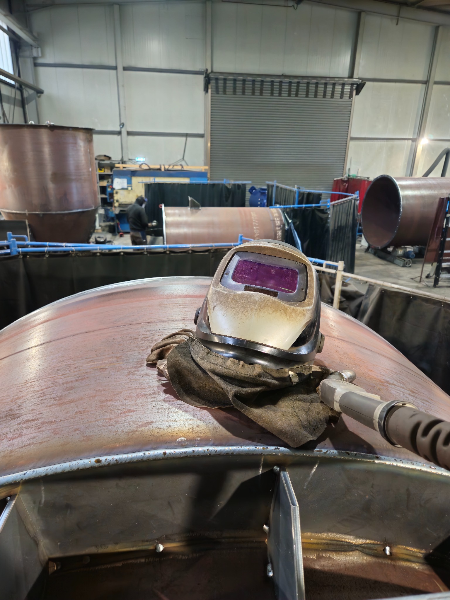 Hełm spawalniczy i szlifierka na metalowej konstrukcji w hali produkcyjnej. W tle widoczny spawacz przy pracy i elementy konstrukcji stalowych.