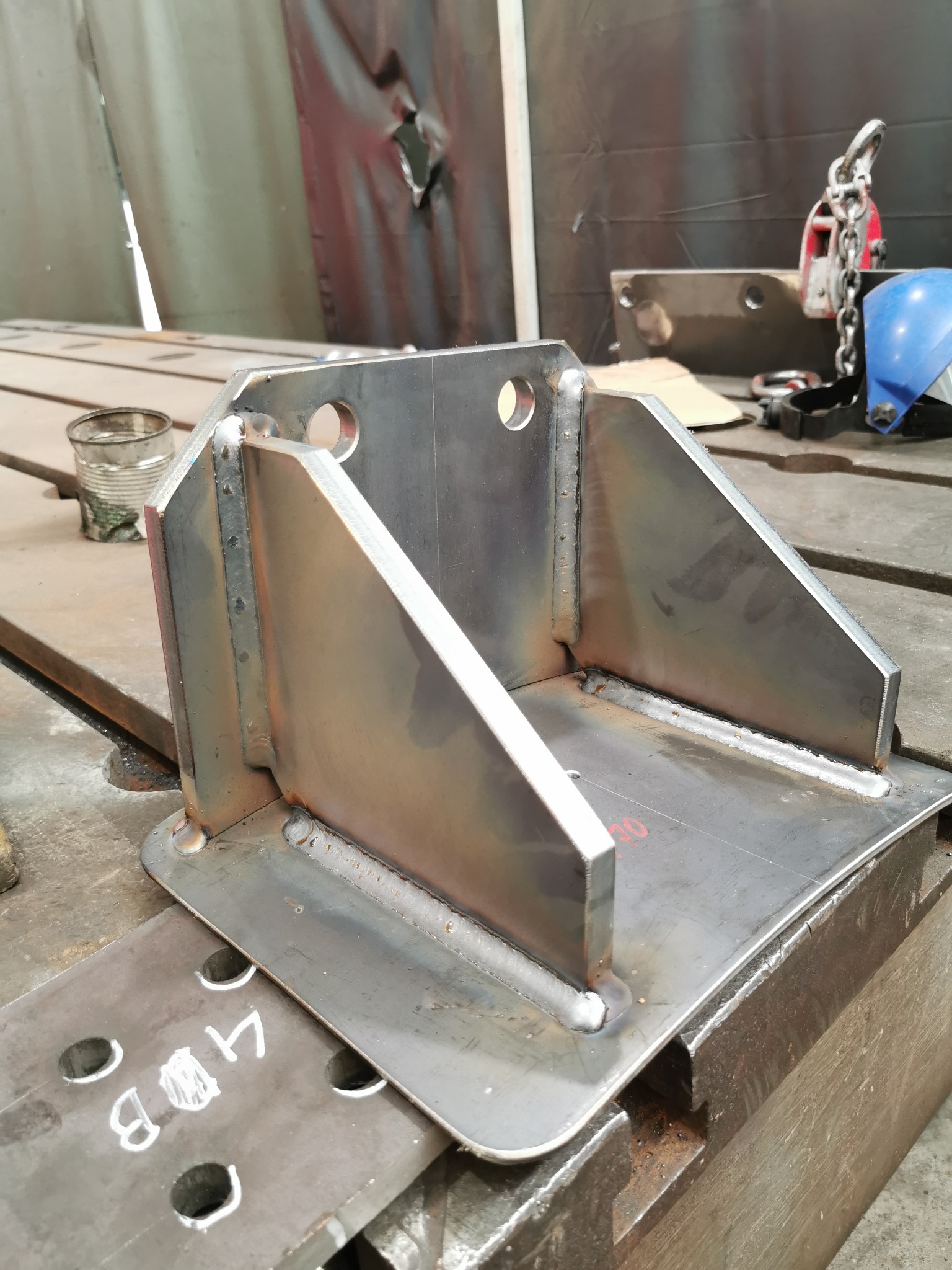 Metalowa konstrukcja spawana z otworami, widoczne spawy, leżąca na stole warsztatowym, w tle narzędzia i akcesoria spawalnicze. Detal konstrukcyjny.