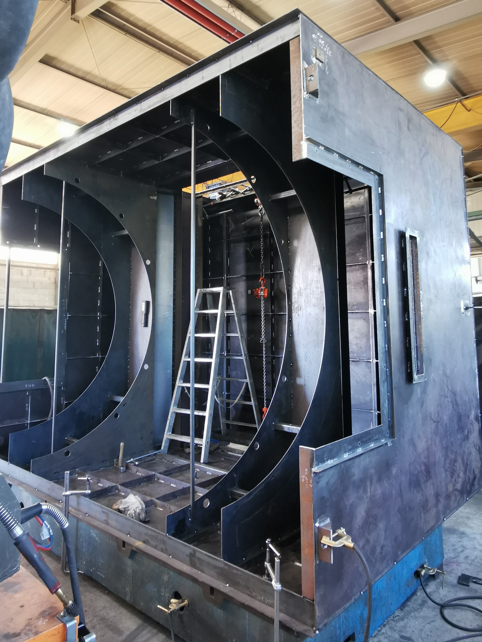 Konstrukcja stalowa w hali produkcyjnej, widoczne elementy spawane i drabina. Duży, przestrzenny szkielet z metalu, etap budowy, surowe wykończenie.