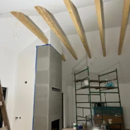 Wnętrze w trakcie wykańczania: szary kominek z maskującą taśmą, drewniane belki na suficie, rusztowanie malarskie z farbami, widoczne przewody elektryczne.