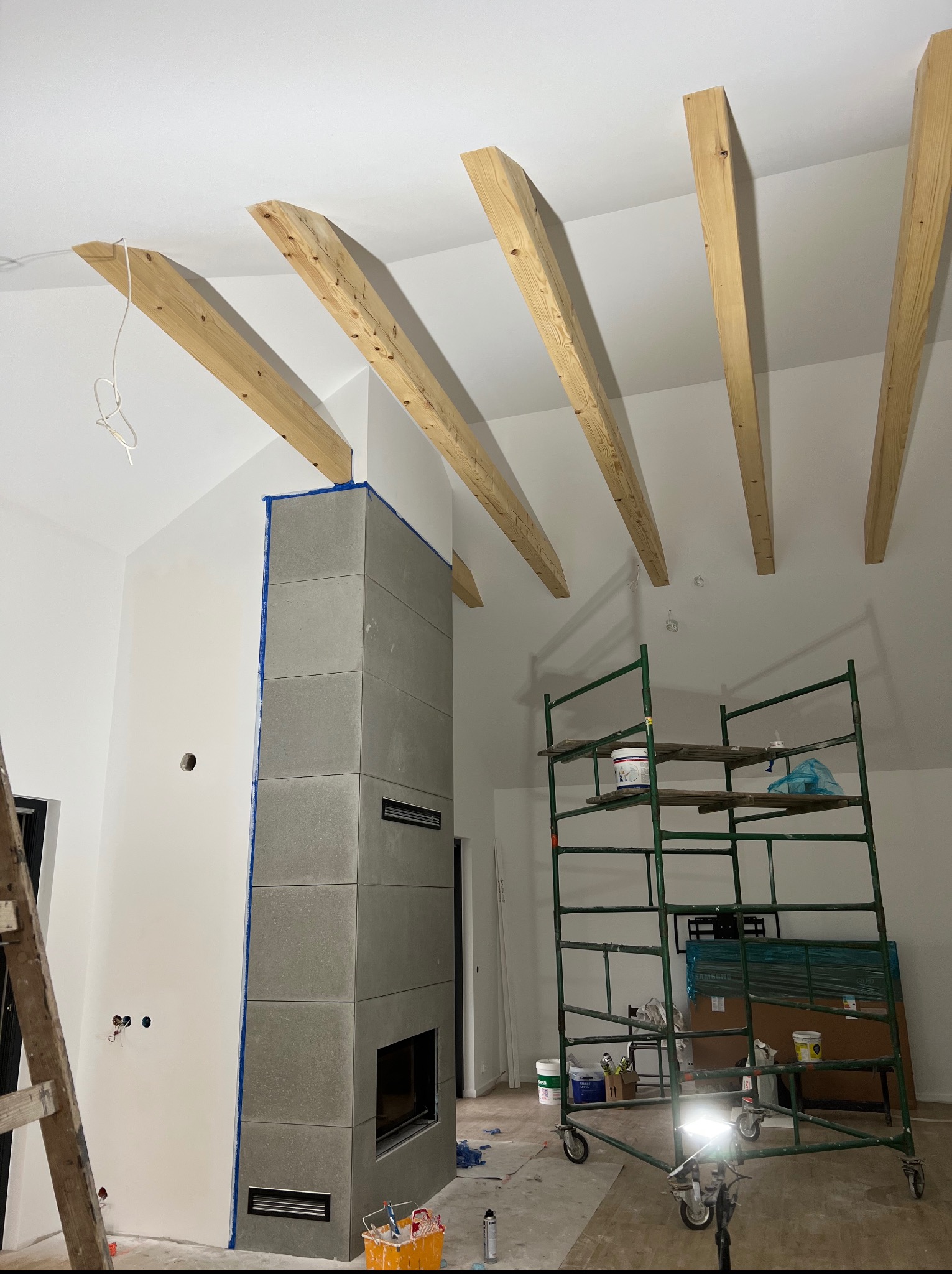 Wnętrze w trakcie wykańczania: szary kominek z maskującą taśmą, drewniane belki na suficie, rusztowanie malarskie z farbami, widoczne przewody elektryczne.
